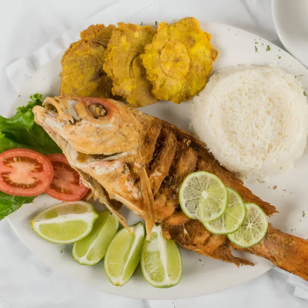 A Colombian fried fish dish with rice, plantains, lime, lettuce, and tomatoes on a white plate.