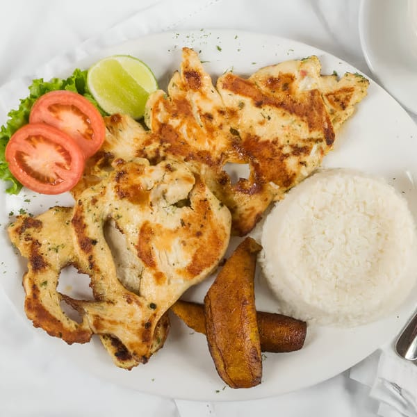 Grilled chicken with rice, fried plantains, lettuce, tomato, and lime on a white plate.