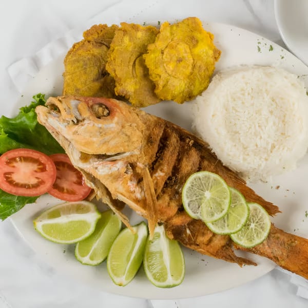 A whole fried fish with lime, rice, plantains, lettuce, and tomato slices on a white plate.