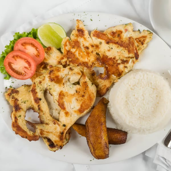 Grilled chicken with rice, fried plantains, lettuce, tomato slices, and a lime wedge on a white plate.