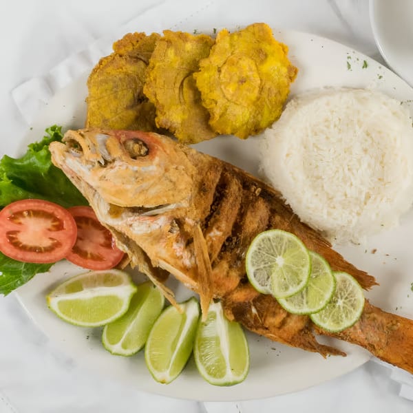 Fried whole fish with lime, rice, plantains, lettuce, and tomato on a white plate.