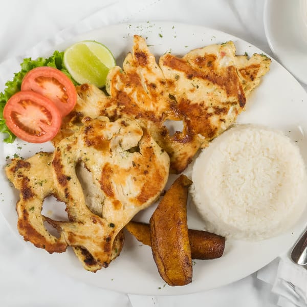 Grilled chicken with rice, fried plantains, lettuce, tomato slices, and a lime wedge on a white plate.