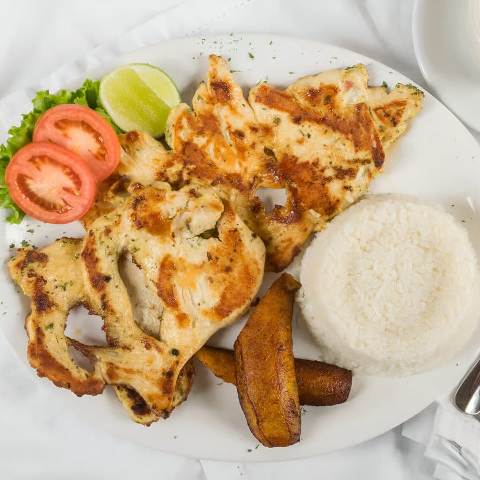 Grilled chicken with rice, fried plantains, lettuce, tomato slices, and a lime wedge on a white plate.