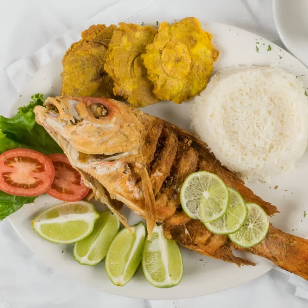 Whole fried fish with limes, rice, tostones, lettuce, and sliced tomatoes on a white plate.