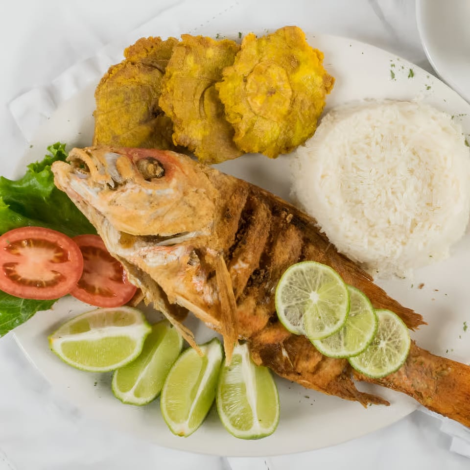 Whole fried fish with limes, rice, tostones, lettuce, and sliced tomatoes on a white plate.