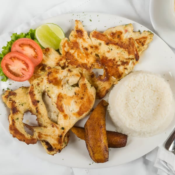 Grilled chicken with rice, fried plantains, lettuce, tomato, and lime on a white plate.